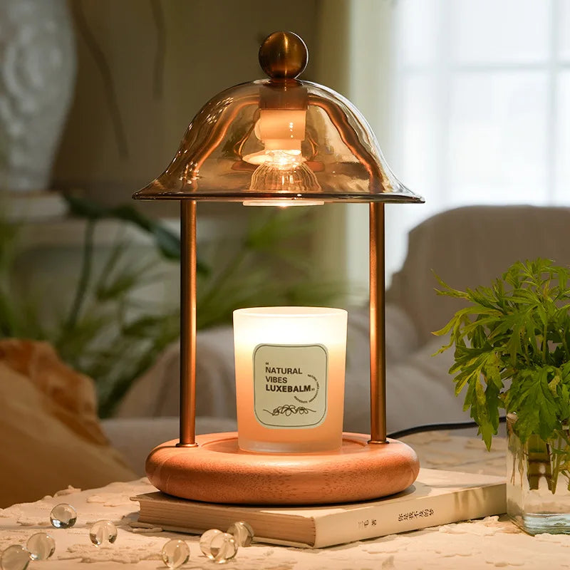 Candle in a wooden holder with a glass cover on a table, surrounded by plants and books.