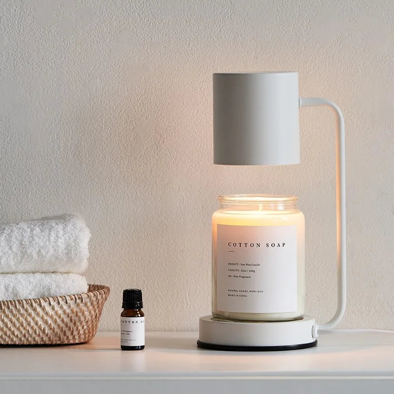 Candle with a diffuser on a surface next to a basket of towels and a bottle, against a neutral wall.